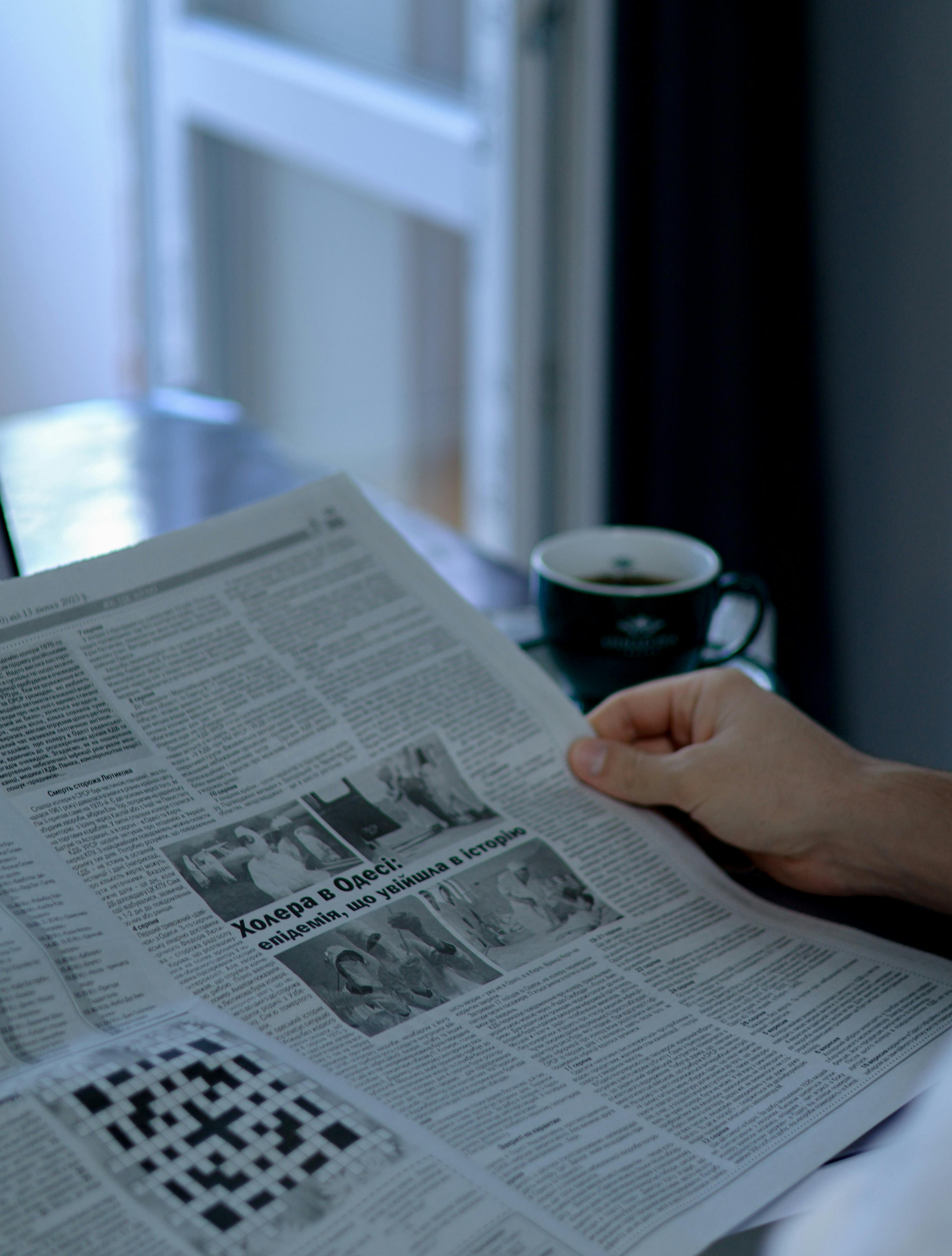 Picture of a hand holding a newspaper with a cup of coffee beside it